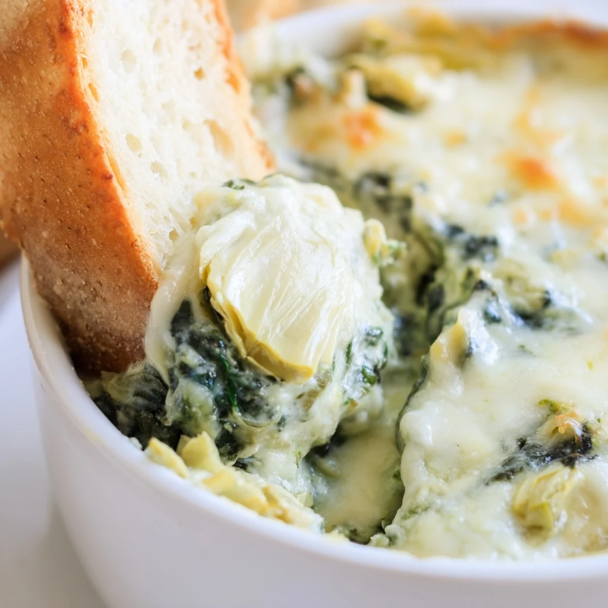 A close-up of creamy spinach and artichoke dip in a white baking dish, with golden, bubbly cheese on top and slices of toasted baguette ready for dipping.  