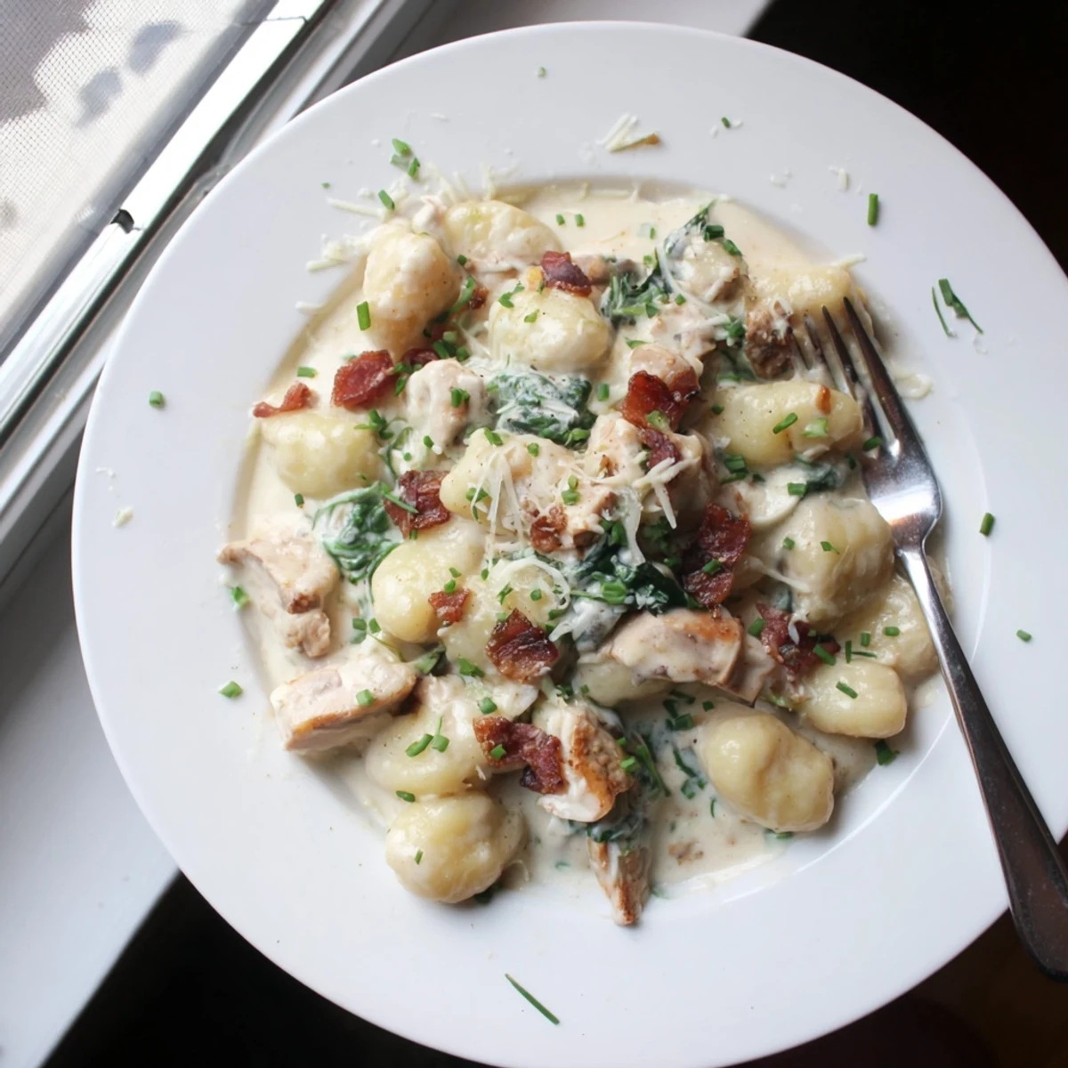 Serving of cheesy chicken gnocchi topped with bacon and fresh chives on a white plate